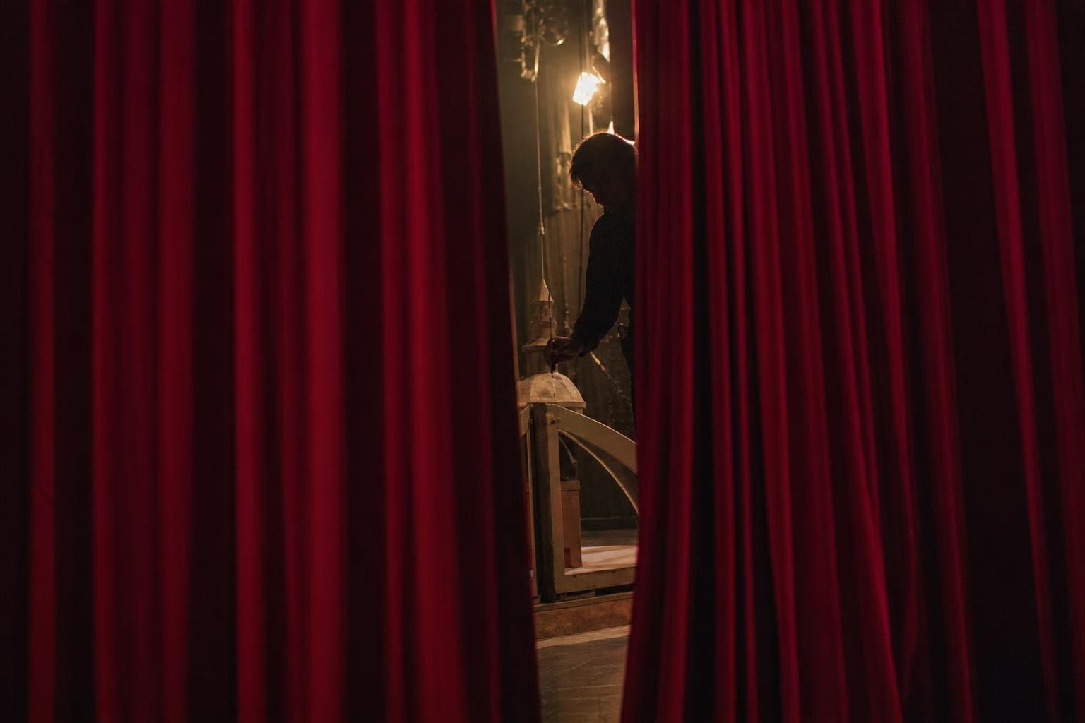A theater curtain closed during rehearsal, with a stagehand barely visible through a narrow gap.