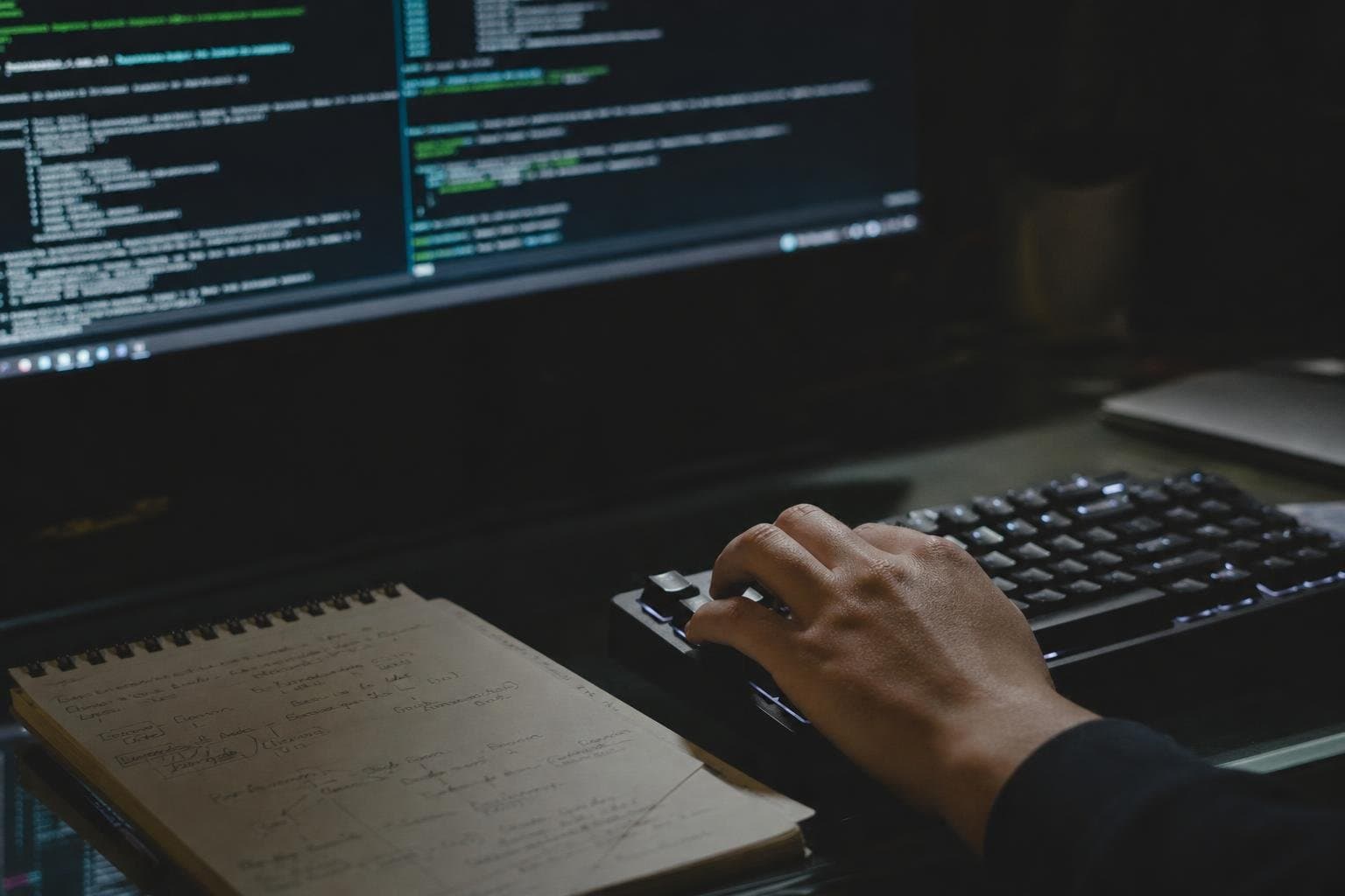 A developer typing at a keyboard at night with a handwritten notepad beside them.
