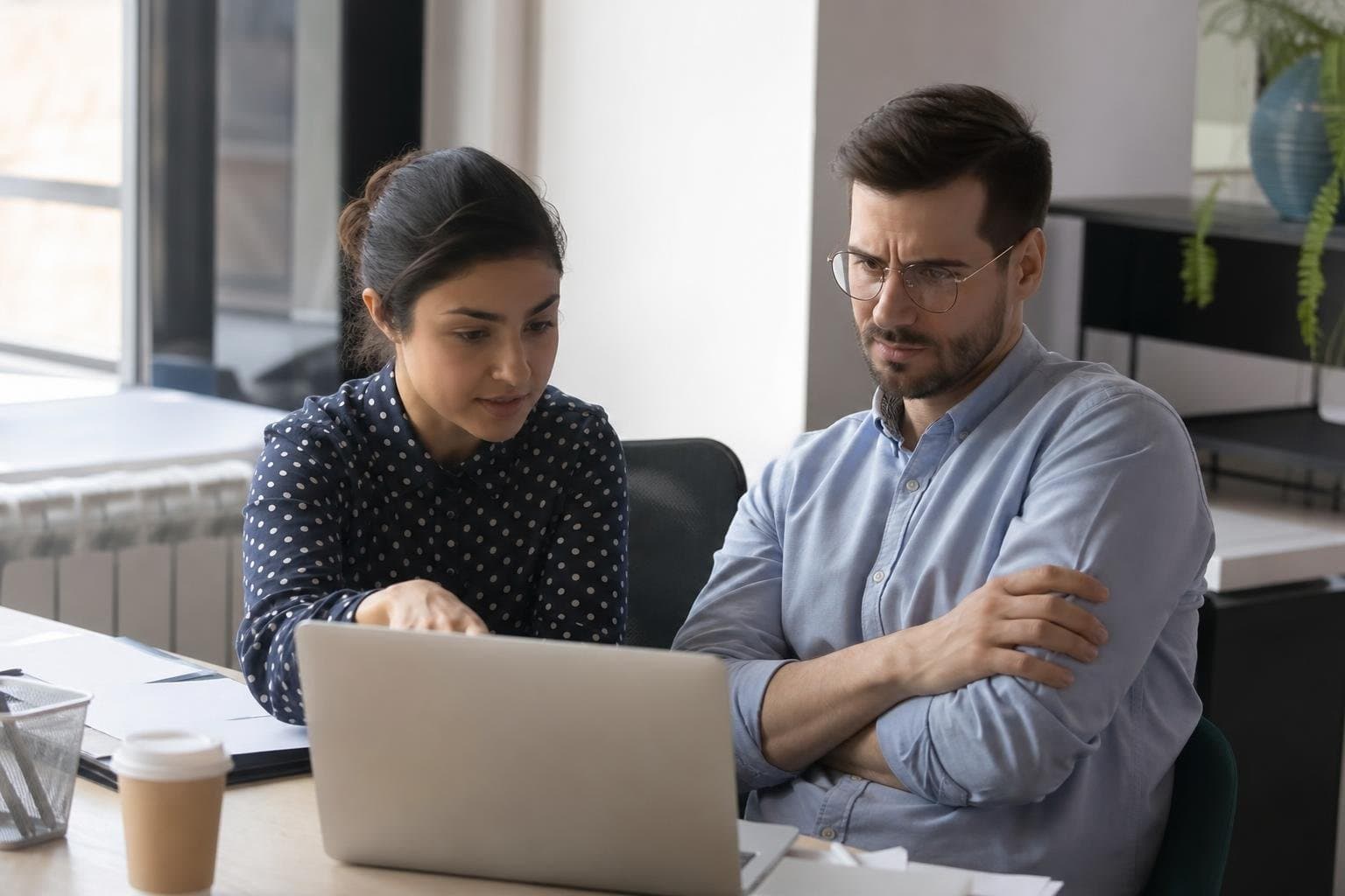 Two colleagues looking at a laptop screen together, one pointing, the other unsure.