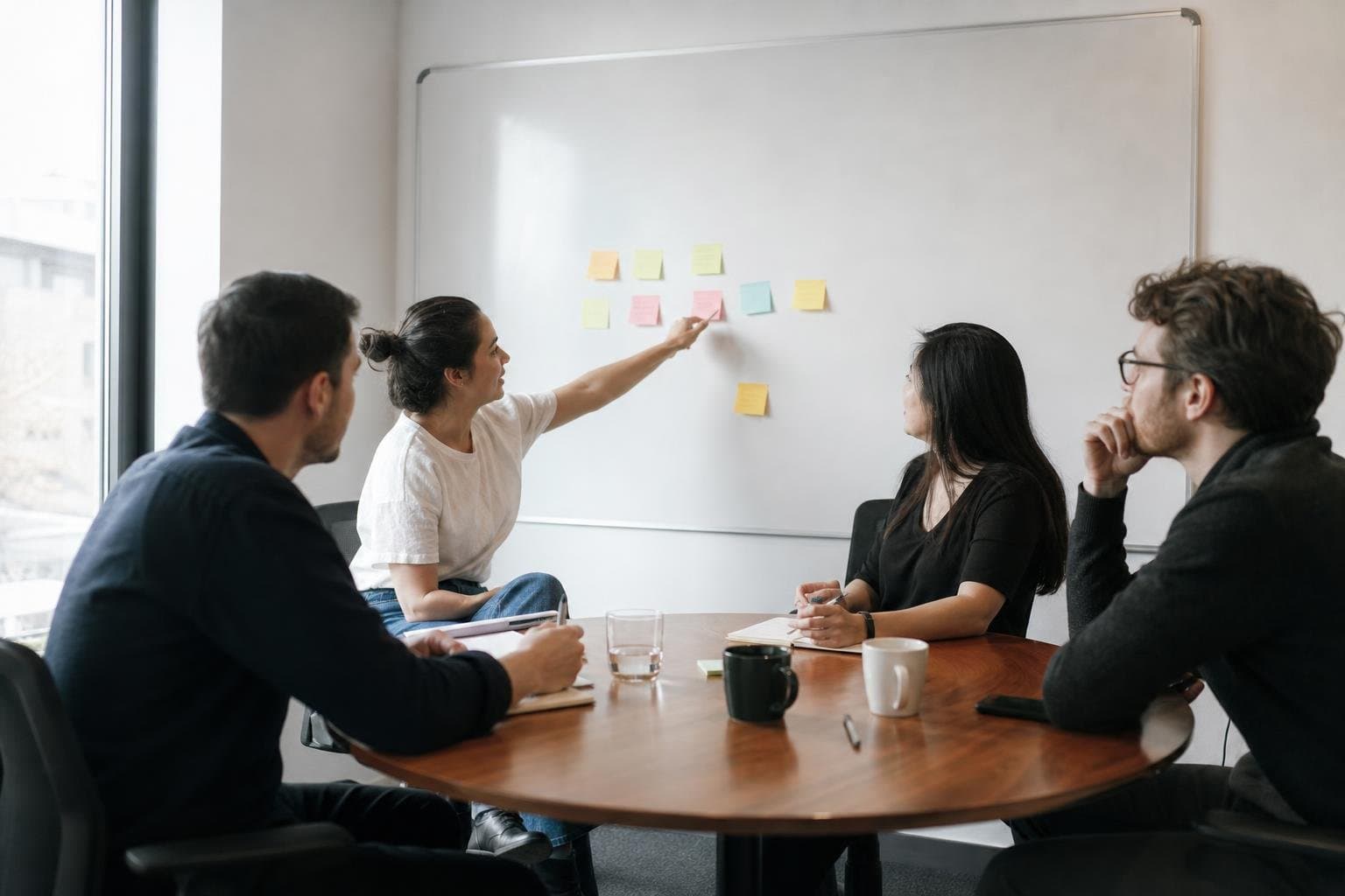 Four colleagues in a small meeting room discussing project status around a whiteboard.
