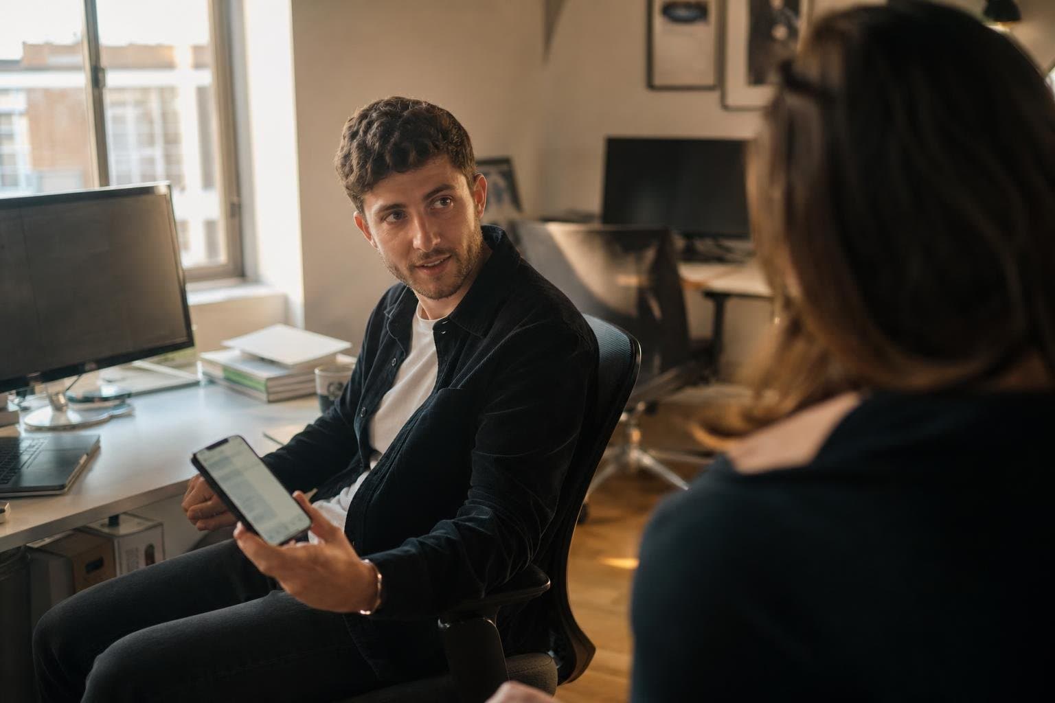 A designer turning to a colleague, phone in hand, in a small sunlit office.